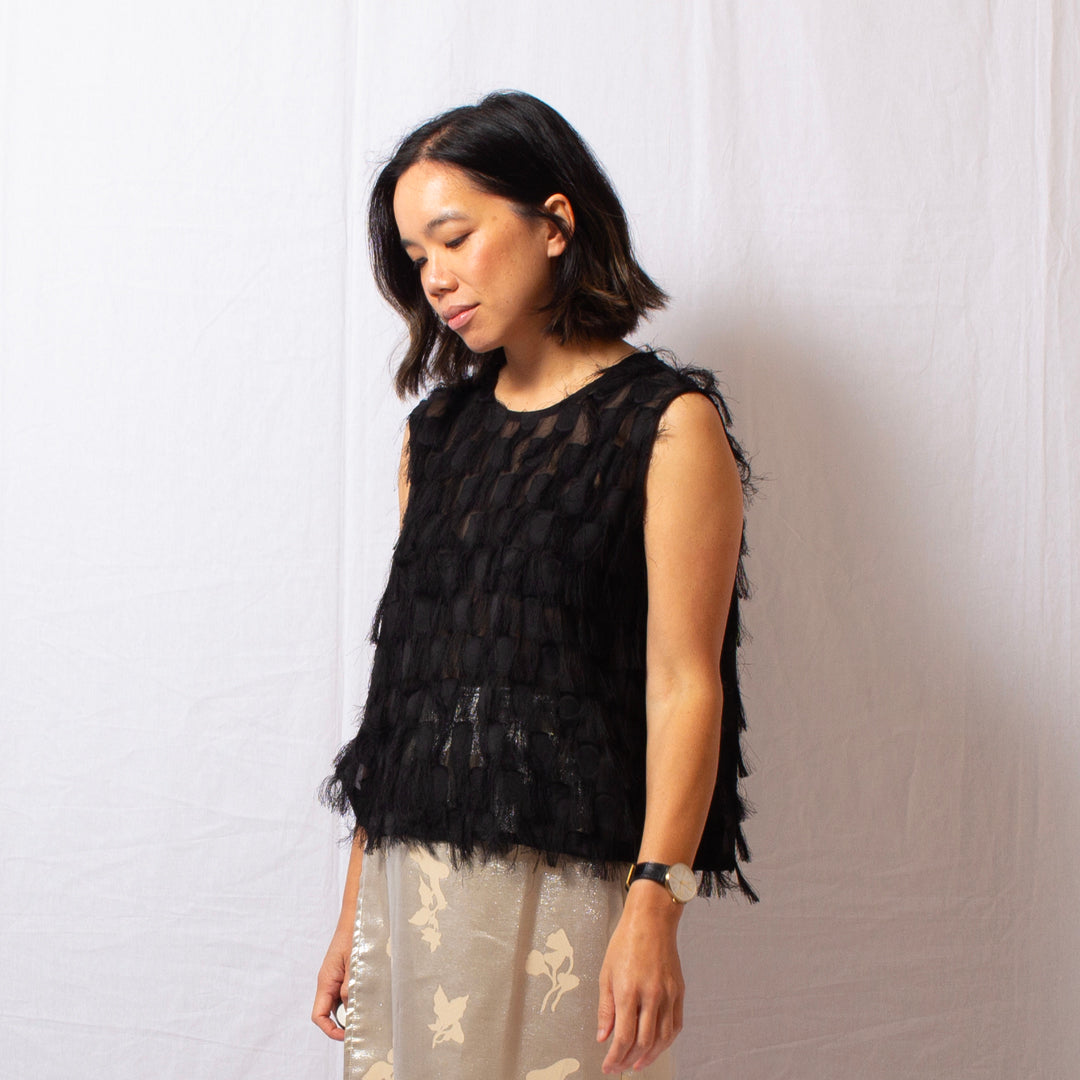 Woman wears black sleeveless fringe top that is semi-sheer with round high neckline, worn with silk skirt.