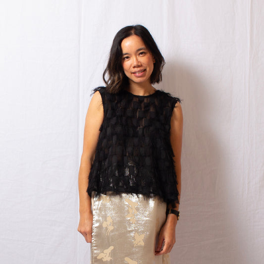 Woman wears black sleeveless fringe top that is semi-sheer with round high neckline, worn with silk skirt.