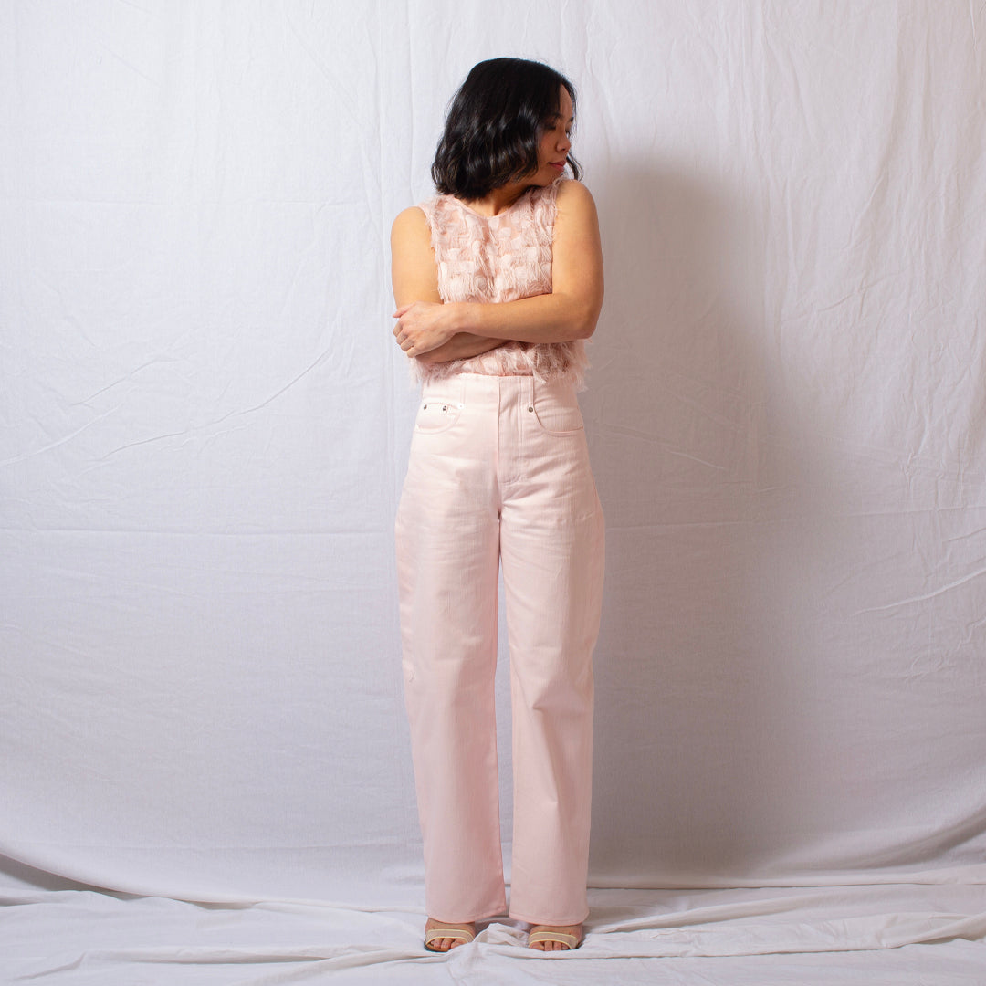 A woman stands with her back to the camera wearing a light pink sleeveless top and matching light pink wide-leg jeans, posed against a wrinkled white fabric backdrop.