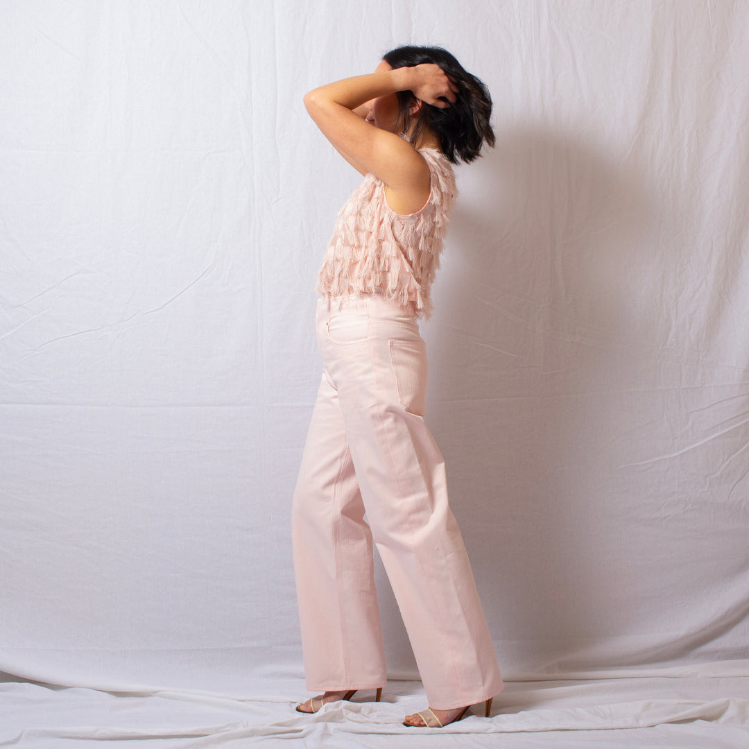 A woman stands in profile wearing a pale blush pink sleeveless top with layered ruffles and matching wide-leg trousers, posed against a white fabric backdrop.