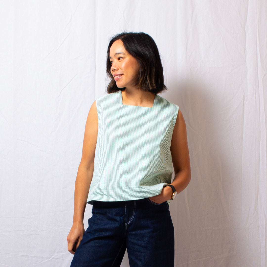 Woman wearing a green and white vertical stripe sleeveless top made from a textured cotton blend seersucker, that has a square high neckline and a relaxed fit, with dark blue jeans.