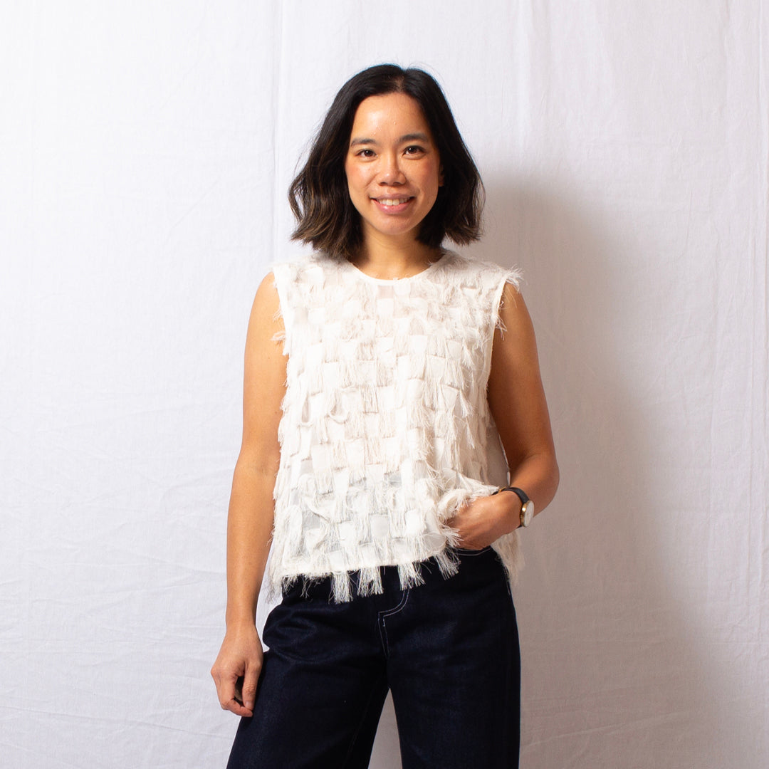 Woman wears ivory sleeveless fringe top that is semi-sheer with round high neckline, worn with navy blue jeans.