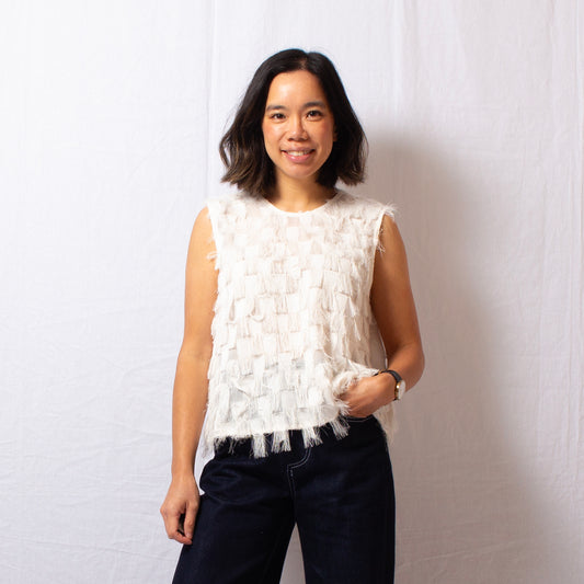 Woman wears ivory sleeveless fringe top that is semi-sheer with round high neckline, worn with navy blue jeans.
