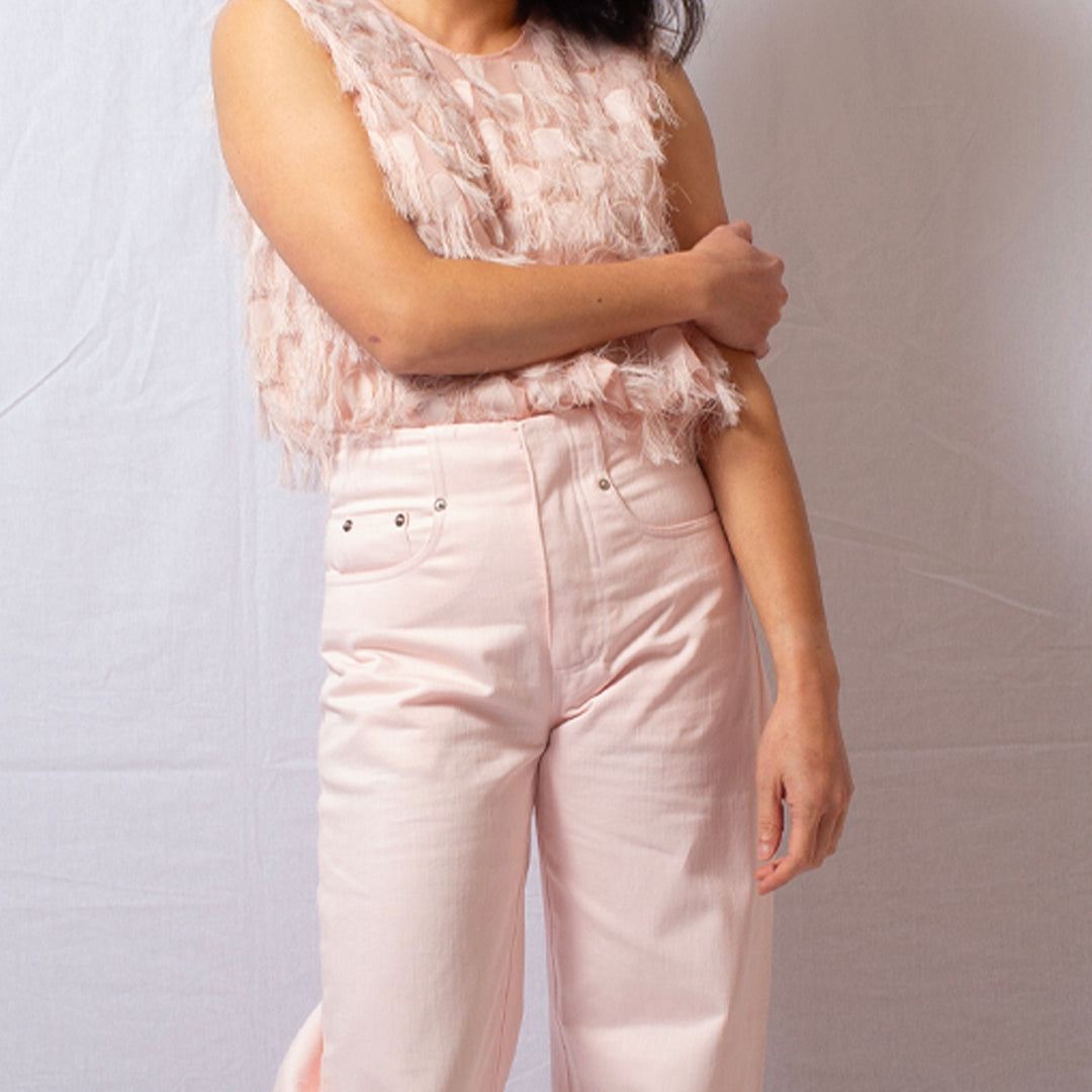 A person wearing a sleeveless light pink feathered top and pale pink high-waisted jeans, standing against a light gray fabric backdrop.