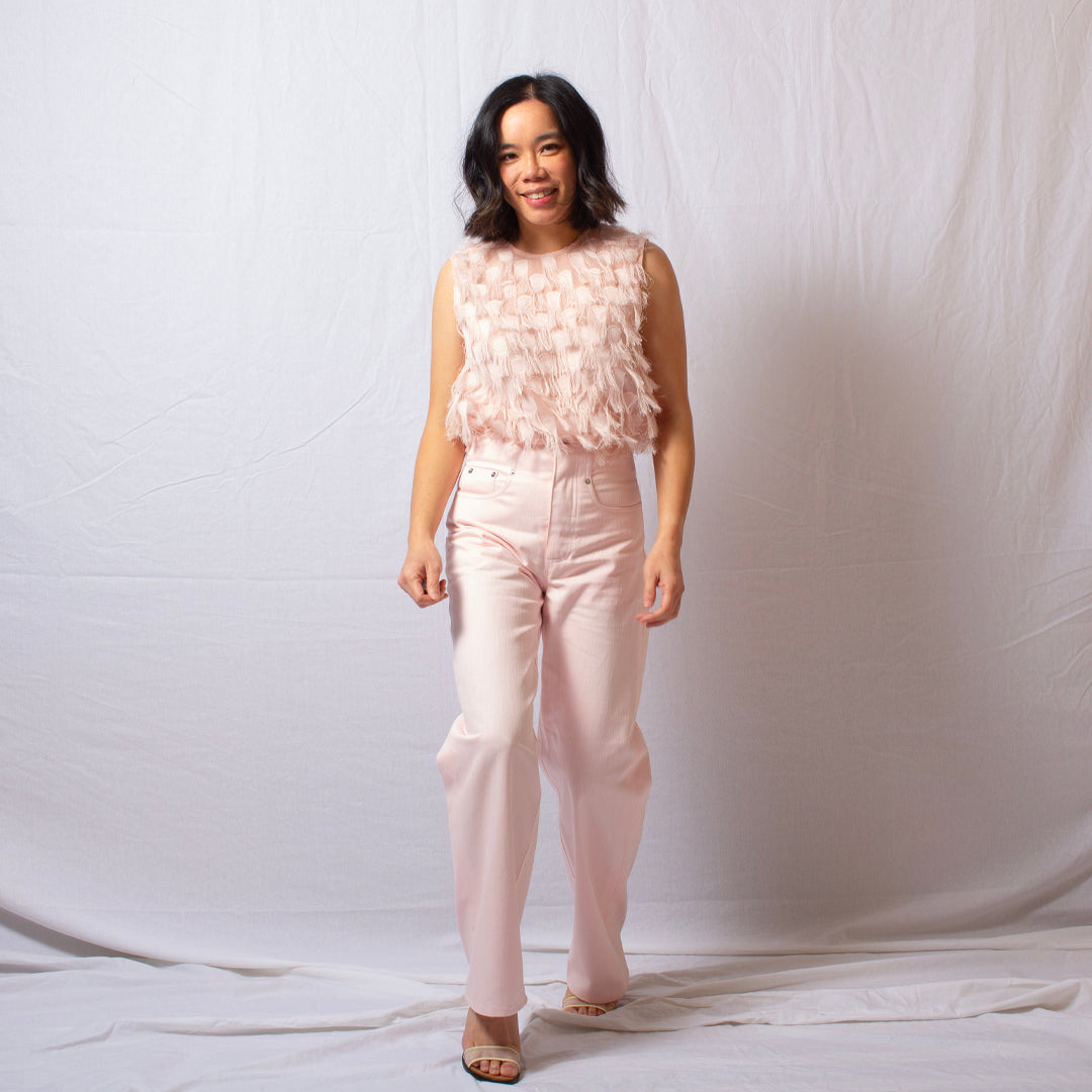 A woman modeling a light pink sleeveless top with a textured, feather-like ruffle overlay and matching light pink high-waisted straight-leg jeans, standing against a white fabric backdrop.